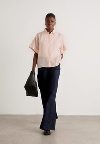 Camicia button-up rosa chiaro a maniche corte, vestibilità comoda, abbinata a jeans denim scuri a zampa. La modella porta una borsa nera. Sfondo neutro.