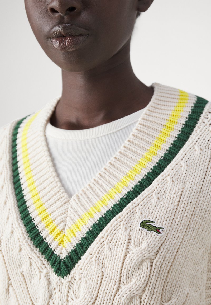 Pull en laine torsadée de couleur crème, avec un col en V bordé de rayures vertes et jaunes et un petit logo de crocodile brodé sur la poitrine.