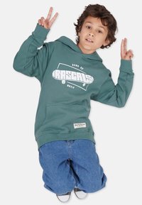 Grüne Kapuzenjacke mit weißem Grafiktext "BAND OF RASCALS" und einem Skateboard-Design. Kombiniert mit blauen lässigen Jeans und einem Markenakzent auf der Kapuzenjacke.