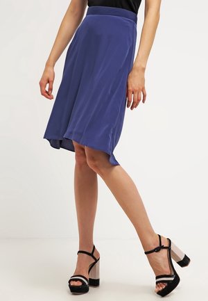 Frau trägt einen blauen asymmetrischen Rock und schwarze Sandalen mit hohem Blockabsatz, steht auf einem weißen Boden mit einfachem Hintergrund.