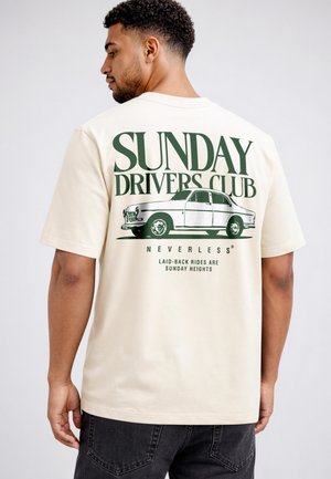 Mann trägt beiges T-Shirt mit Vintage-Auto-Grafik und Text „Sunday Drivers Club“ und „Laid-back rides are Sunday heights“ auf dem Rücken.