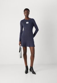 Marineblauwe geribbelde langejurk met een aansluitende silhoutte en een uitlopende zoom. Gepaard met zwarte puntige enkelboots en een zilveren handtas.