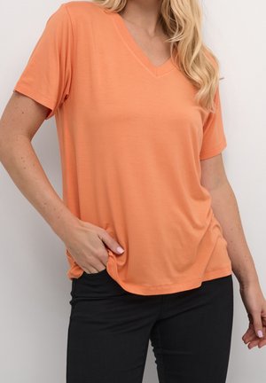Femme portant un T-shirt orange à manches courtes et col en V, ample, avec un pantalon noir, main posée dans la poche, sur fond blanc.