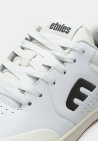 Λευκό αθλητικό παπούτσι με υφή από δερμάτινο επάνω μέρος, με μαύρες λεπτομέρειες στο λογότυπο, επίπεδες λευκές κορδόνες και τρυπητό μπροστινό τμήμα.