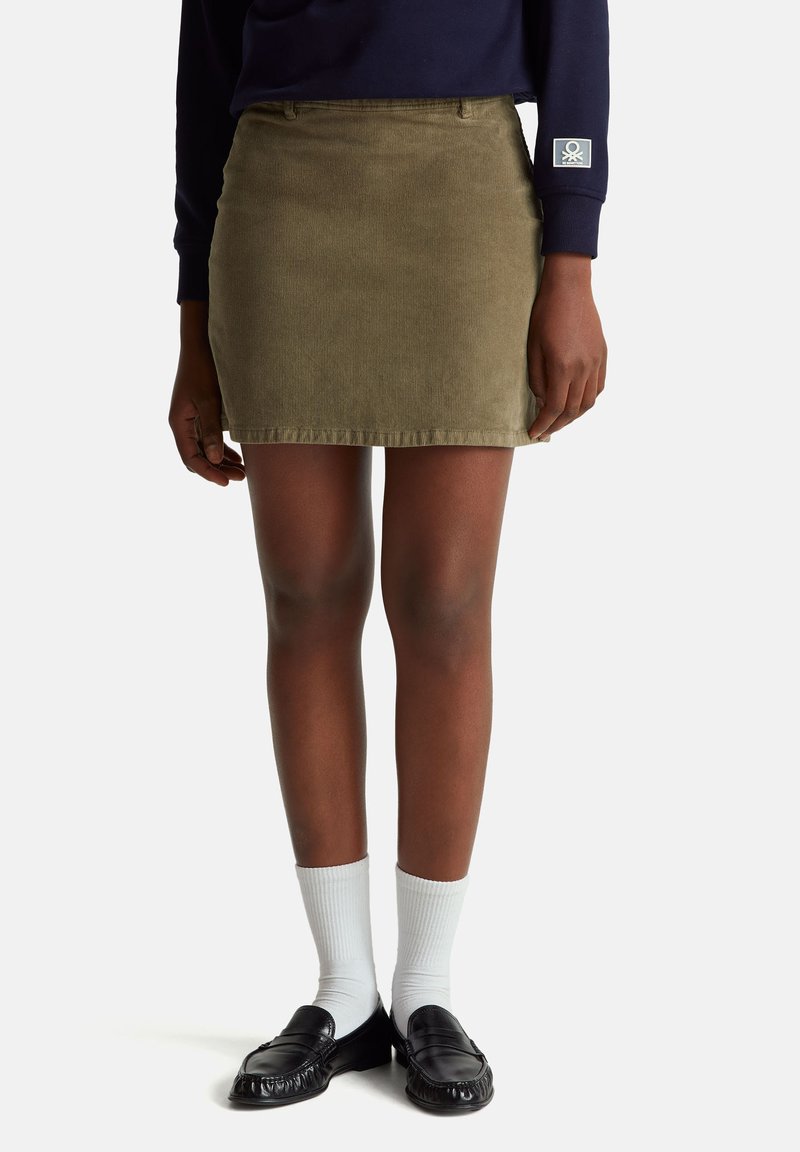 Olivově zelená midi sukně z kordu s texturovaným povrchem, předními kapsami a přiléhavým střihem. Spojena s černými mokasíny a bílými ponožkami.