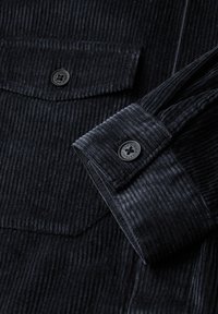 Černá corduroy košile s texturovaným povrchem, dvěma náprsními kapsami a kulatými černými knoflíky s květinovým vzorem.