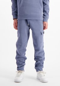 Persoon in blauwe joggingbroek met zijzakken en witte sneakers, staand tegen een witte achtergrond.
