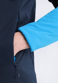 Navyjacka med geometrisk textur, med en kontrasterande blå ärm. handen når ner i en dragkedjeficka på sidan.