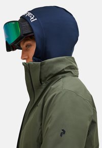 Person in einer marineblauen Kapuze, Skibrille mit grünen, reflektierenden Gläsern und einer olivgrünen, hochkragen Jacke, der nach links zeigt, vor einem weißen Hintergrund.