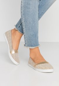Des chaussures slip-on tissées beiges avec des accents dorés, une semelle blanche et un bout arrondi. Portées avec un jean frangé bleu clair, mettant en valeur un style décontracté.