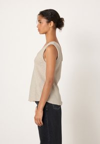 Keskmise tumeda nahatooniga naine, kellel on lokkis juuksed kuklas, kannab varrukateta beežikat toppi ja tumedaid kõrge vöökohaga teksaseid ning seisab profiilis valge tausta ees.