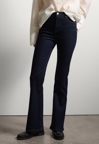 Jeans svasati blu scuro in denim, con vita alta, cuciture a contrasto e hardware minimale, abbinati a mocassini neri.