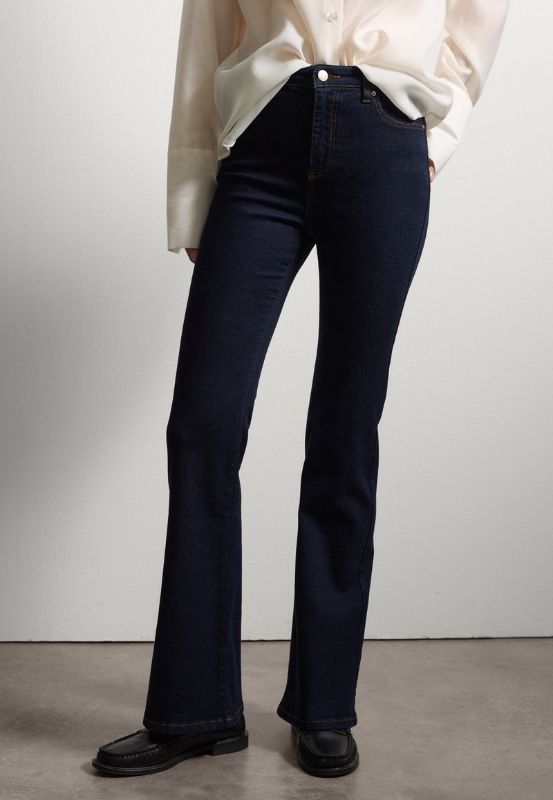 Jeans svasati blu scuro in denim, con vita alta, cuciture a contrasto e hardware minimale, abbinati a mocassini neri.