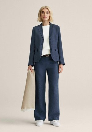 Lyshåret kvinde iført navy jakkesæt med hvid sweater og sneakers holder beige tørklæde og står mod en ensfarvet lys baggrund.