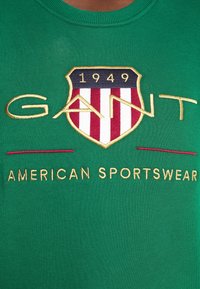 Groene katoenen T-shirt met een geborduurd schild met rode, witte en blauwe strepen. Tekst leest "GANT" en "AMERICAN SPORTSWEAR" in goud.