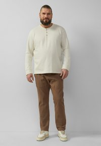 Beige langärmeliges henley-shirt met drie knopen, gecombineerd met een bruine broek en witte sneakers, tegen een lichte achtergrond.