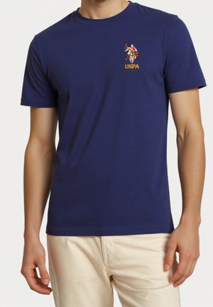 Homme portant un t-shirt bleu marine avec un petit logo multicolore joueur de polo et le texte "USPA" sur la poitrine, assorti à un pantalon beige.