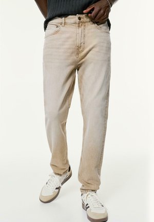 Beige Denim-Jeans mit klassischem gerade geschnittenem Design, fünf Taschen und Knopfverschluss. Kombiniert mit weißen und grauen Turnschuhen.