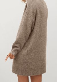 Robe pull en tricot beige avec une coupe décontractée, des manches longues, des poignets et un ourlet côtelés, et une texture douce. Aucun motif distinct ou élément en métal visible.