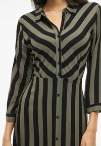 Šaty s gombíkmi vyrobené z hladkej látky, so zvislými olivovo zelenými a čiernymi pruhmi, s golierom a manžetami na trojštvrťové rukávy.