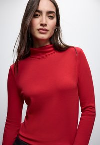 Pull en maille rouge à col roulé et manches longues, fabriqué dans un tissu doux et lisse. Présente un col haut et une silhouette ajustée, complétés par un design minimaliste.
