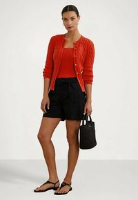 Cardigan de tricô canelado vermelho sobre uma blusa combinando, calções pretos de cintura alta, bolsa tecida preta, sandálias baixas, fundo neutro.