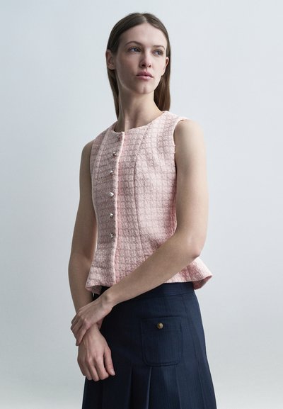 Jeune femme aux cheveux raides portant un haut sans manches rose texturé à boutons et une jupe bleu foncé avec poche, debout devant un fond clair.