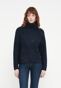 Pull en maille bleu marine avec col montant, doté de motifs en diamant et orné de brillants accents métalliques éparpillés. Tissu souple et texturé.