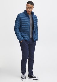 Blauer Puffer-Jacke mit horizontalen Nähten, grauem Pullover, marineblauen Chinos und schwarzen Sneakers. Die Jacke verfügt über einen Reißverschluss vorne und einen hohen Kragen.