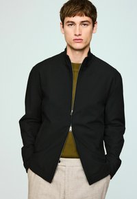 Črno suknjič z zadrgo in visokim ovratnikom, z dvema stranskima žepoma, oblečen čez olivno zelen pulover. Gladek material z minimalnim šivanjem.