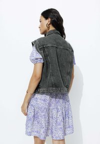 Gilet en denim noir à manches retroussées sur une robe courte à motifs avec des pois bleus sur un fond clair. Détails de design et de texture simples visibles.
