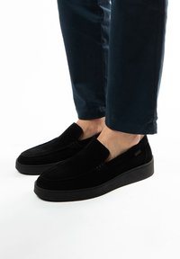 Black suede slip-on shoes with a flat, black rubber sole, featuring minimalistic design and no visible hardware or embellishments.