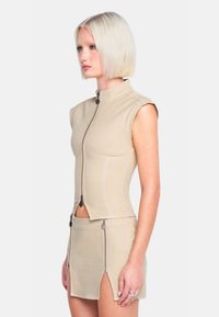 Beige ärmelloses Oberteil mit hohem Kragen und Frontreißverschluss, kombiniert mit einem passenden Rock, der Reißverschlüsse und seitliche Schlitze aufweist. Glatte Textur.