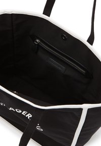 Sac fourre-tout en tissu noir avec des bords blancs, doté d'une poche intérieure zippée et d'un détail de logo blanc à l'extérieur. Texture douce.
