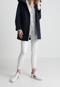 Parka con capucha en color navy, camisa de botones a rayas, jeans ajustados blancos, zapatillas blancas con acentos negros, tejido texturizado, diseño casual.
