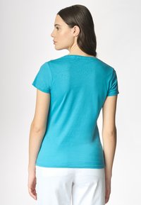 Una donna con lunghi capelli castani indossa una maglietta azzurro turchese a maniche corte, girata di spalle contro uno sfondo chiaro.