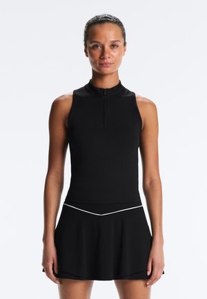 Žena s vlasmi stiahnutými dozadu, oblečená v čiernom tielkovom topi na zips a zodpovedajúcej čiernej sukni s bielym lemovaním, stojaca čelom k bielej stene.