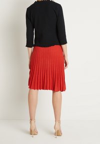 Černý top s krátkým rukávem a volánem v pase, spárovaný s červenou skládací sukní ke kolenům. Outfit doplňují béžové lodičky na kotníkovém pásku.
