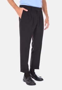 Pantaloni neri su misura con pieghe, chiusura con bottone e orli arrotolati, abbinati a mocassini neri e una camicia azzurra chiara.