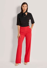 Schwarze Bluse mit V-Ausschnitt und Schleifen-Details, kombiniert mit roten, weit geschnittenen Hosen. Das Model steht vor einem neutralen Hintergrund.