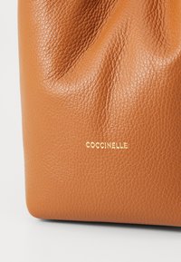 Ruskea teksturoitu nahkakäsilaukku pehmeällä taittosuunnittelulla, jossa on kultainen logo "Coccinelle" lähellä pohjaa.