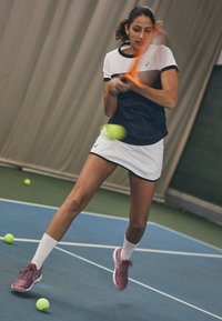 Femme athlétique en tenue de tennis bleu marine et blanche, frappant une balle de tennis jaune avec une raquette orange, portant des chaussures de sport roses.