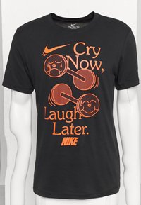 Camiseta de algodón negra con texto naranja: "Llora ahora, ríe después", con gráficos de pesas y caras sonrientes. Logo de Nike en la parte inferior.