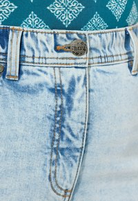 Vaaleansiniset farkut, joissa on haalistunut pinta, kupari-osat, kontrastivärinen ompelu ja napitus vyötäröllä.