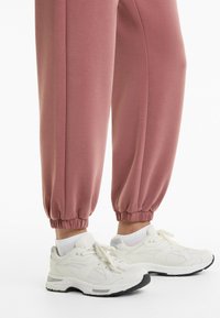 Jalat yang mengenakan celana karet cuff berwarna merah muda debu dipadukan dengan sepatu sneakers atletik putih dan kaus kaki pergelangan kaki putih, berdiri di atas permukaan putih.