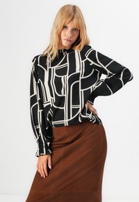 Donna con capelli biondi che indossa una blusa a motivi geometrici in bianco e nero e una gonna marrone, in posa con una mano sulla vita.
