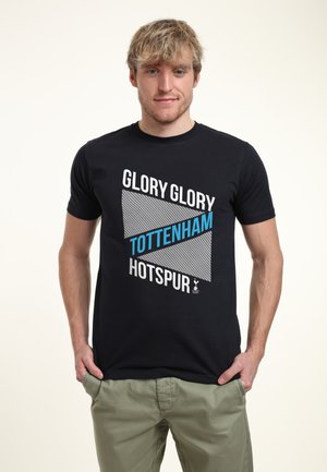 Mladý muž v černém tričku s nápisem "Glory Glory Tottenham Hotspur" a khaki kalhotách, stojící se založenýma rukama v kapsách na jednobarevném pozadí.