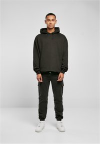 Sudadera negra con cordón ajustable, combinada con pantalones cargo oscuros con bolsillos laterales. El modelo lleva zapatillas blancas. Textura de tela suave.