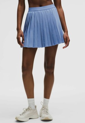 Kvinde iført en lyseblå plisseret mininederdel, hvide ankelsokker og hvide løbesko, stående foran en ensfarvet baggrund.