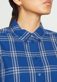 Camisa de cuadros azul con un cuello estructurado, que presenta rayas blancas y azul marino. Tiene botones en un tono de azul ligeramente más oscuro.
